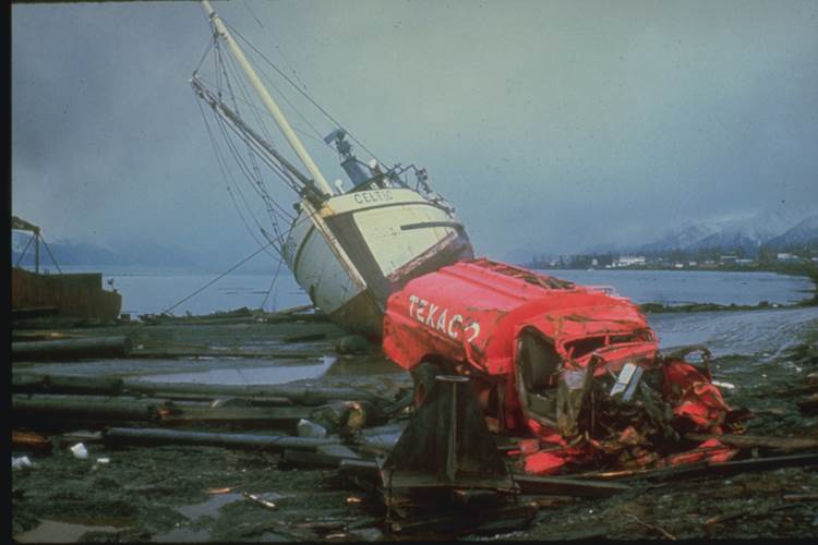 View of the north end of Resurrection Bay at Seward, Alaska, about 75 km from the epicenter. An overturned ship, a demolished Texaco chemical truck, and a torn-up dock strewn with logs and scrap metal are visible. At Seward, a community of about 2,300, a section of the waterfront slid into Resurrection Bay. Waves spread in all directions, destroying the Alaska railroad docks, washing out railroad and highway bridges, and piling railroad rolling stock into giant windrows of wreckage. The waves left a shambles of houses and boats in the lagoon area, some still looking relatively undamaged and some almost completely battered. The waves spread flaming petroleum over the waterfront, igniting the rolling stock, the electrical generation plant, and some residences. Resurrection Bay received $14.6 million in damage. Eleven fatalities occurred in the Seward area. (Source: NOAA/NCEI, US DOI)